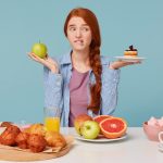 Hard choice. Portrait of beautiful woman trying to choose between healthy and unhealthy food while isolated on bluebackground. Woman holding cake and apple in hands